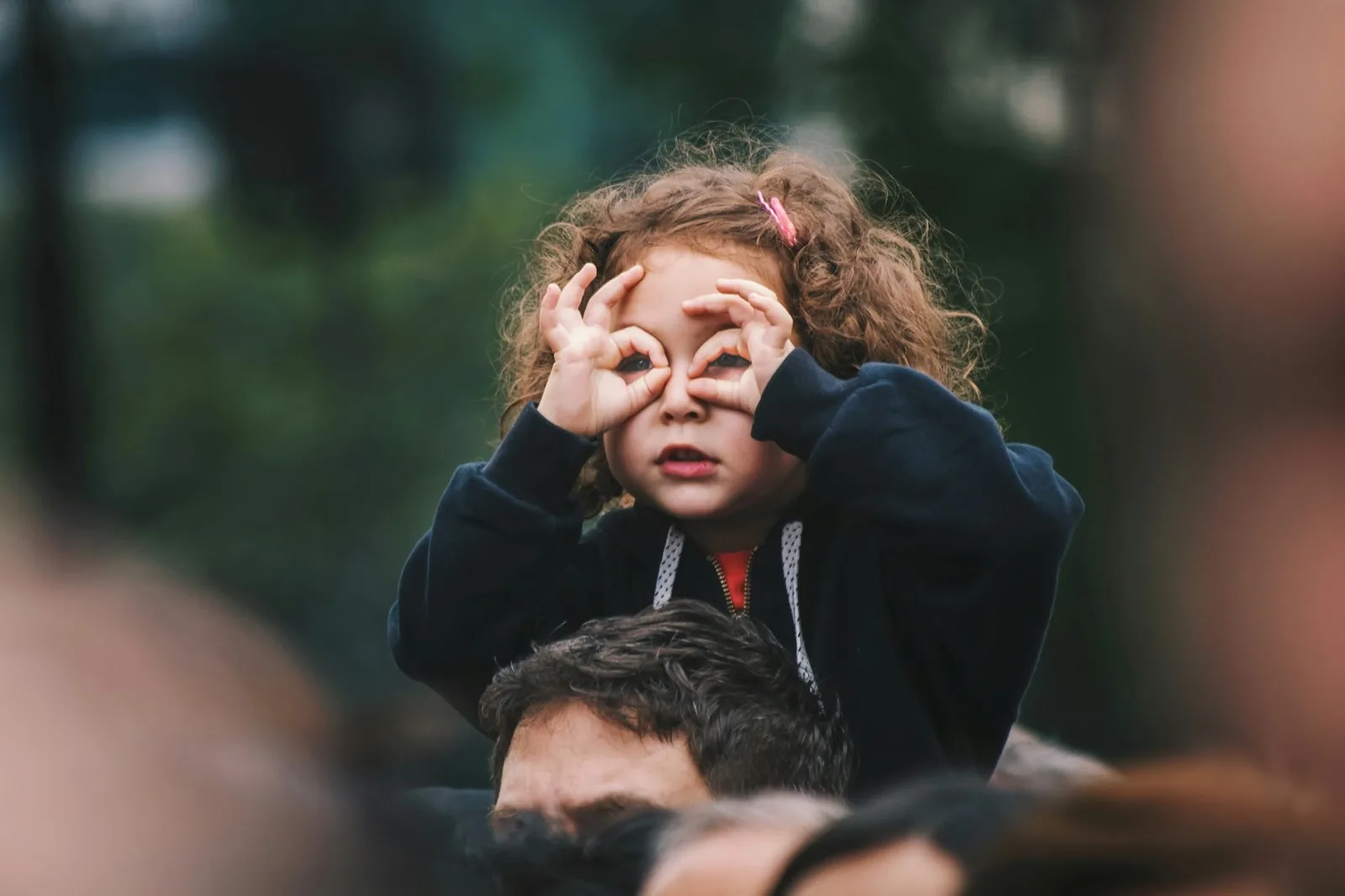 Ein Mädchen auf den Schultern eines Erwachsenen schaut durch eine mit den Händen geformte Brille.
